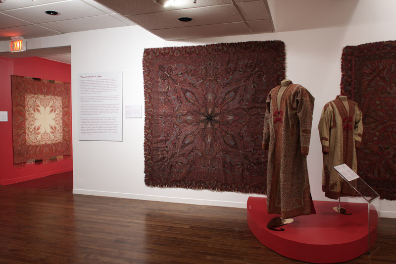 Exhibition with a red and white wall with textiles hanging from the wall and a semicircle red plinth holding two mannequins displaying garments.