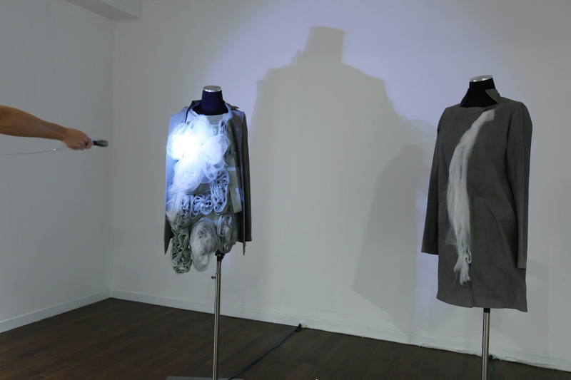 Exhibition with two mannequins standing on the floor displaying garments with a person shining a light on one.