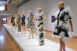 Exhibition with mannequins on a long plinth all facing the same direction displaying Iris Van Herpen garments.