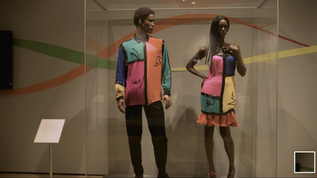 Exhibition with one male and one female mannequins in a glass cabinet displaying garments.