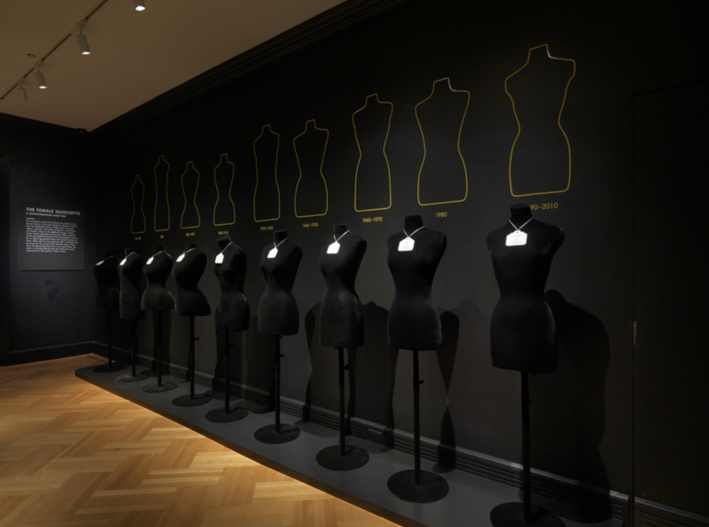 Exhibition with black painted walls and a row of black dressmakers mannequins.