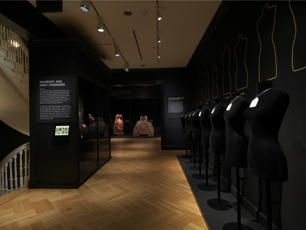 Exhibition with black painted walls and a row of black dressmakers mannequins and text panel and glass cabinet against wall with garment displayed on mannequins.