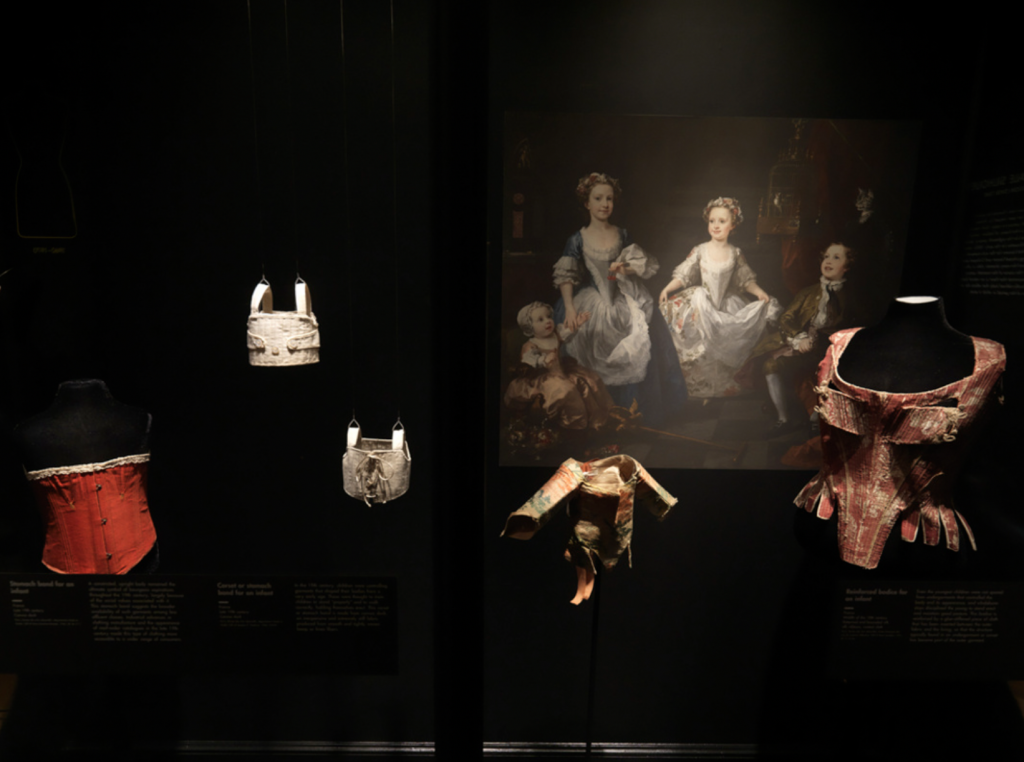 Exhibition with black walls and with glass display cases against the wall with mannequins displaying garments with a print of a painting hanging on the wall.