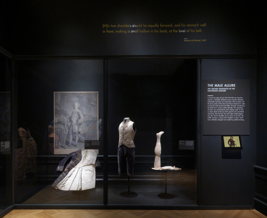Exhibition with black walls and with glass display cases against the wall with mannequins displaying garments.