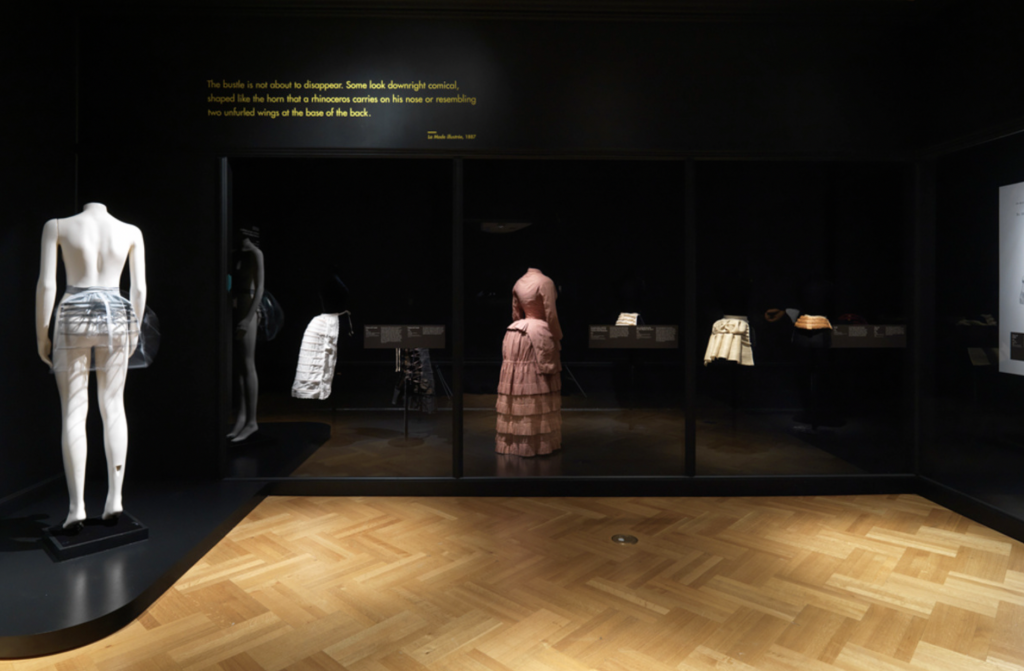 Exhibition with black painted walls and with glass cabinets displaying garments and a mannequin on a plinth.