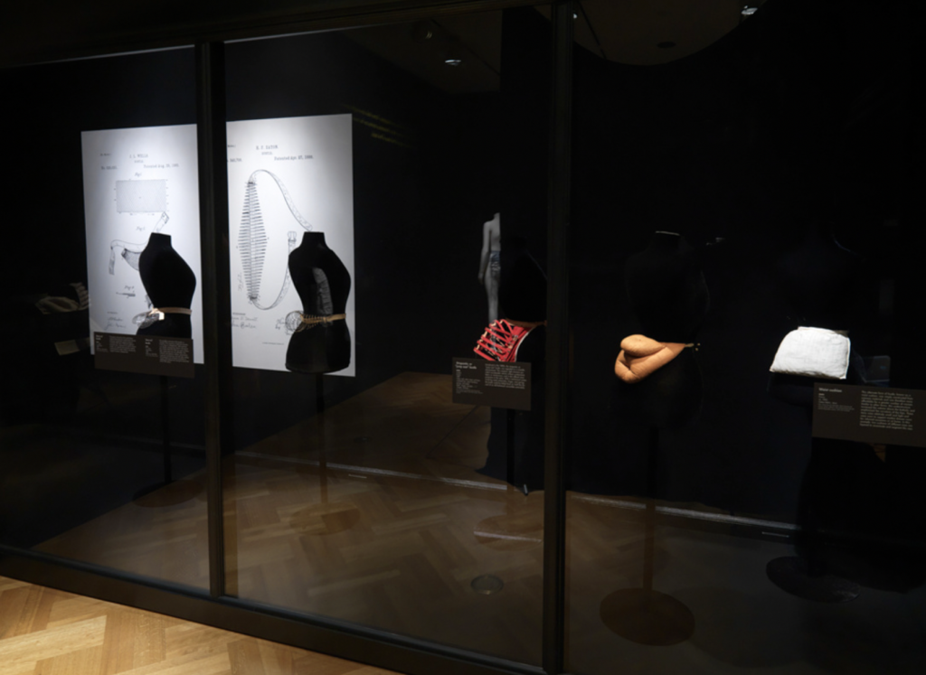 Exhibition with black walls and with glass display cases against the wall with mannequins displaying garments.