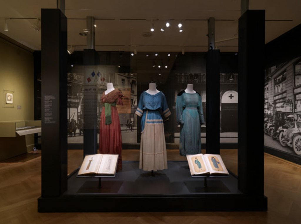 Exhibition with plinth surrounded by glass with three mannequins displaying garments and two open books.