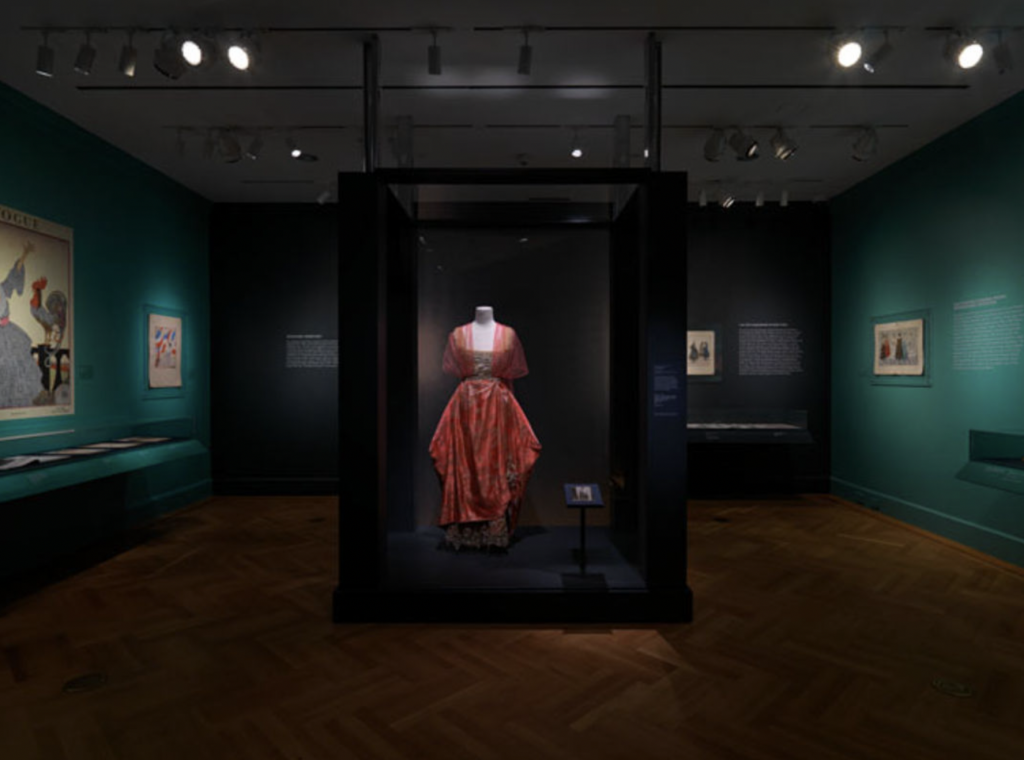 Exhibition with plinth surrounded by glass with a mannequin displaying a dress and photograph.