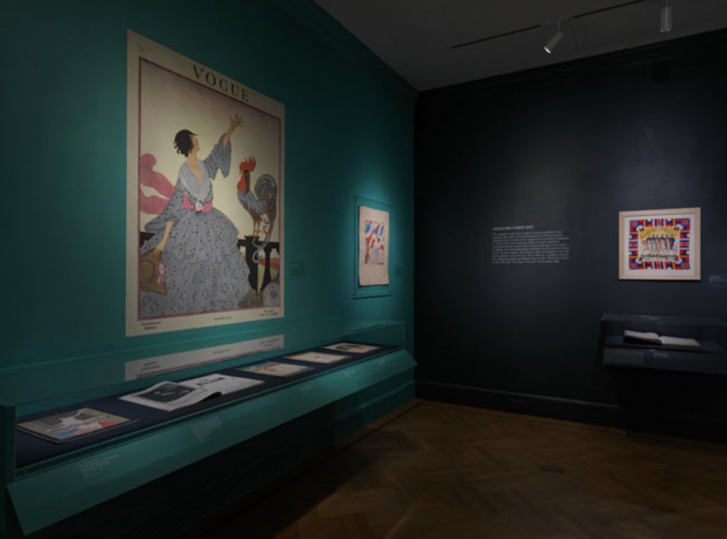 Exhibition with two walls; one turquoise wall with prints hanging and long table height glass cabinet displaying magazines and paper artefacts.