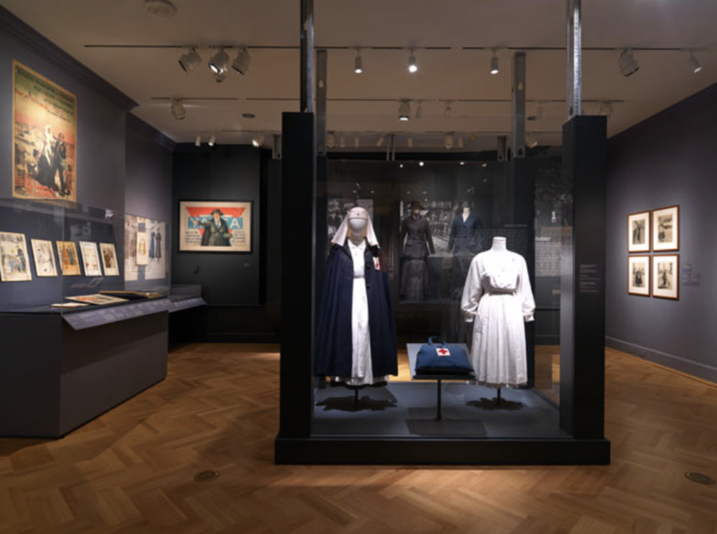 Exhibition with plinth surrounded by glass with two mannequins displaying garments and a bag.