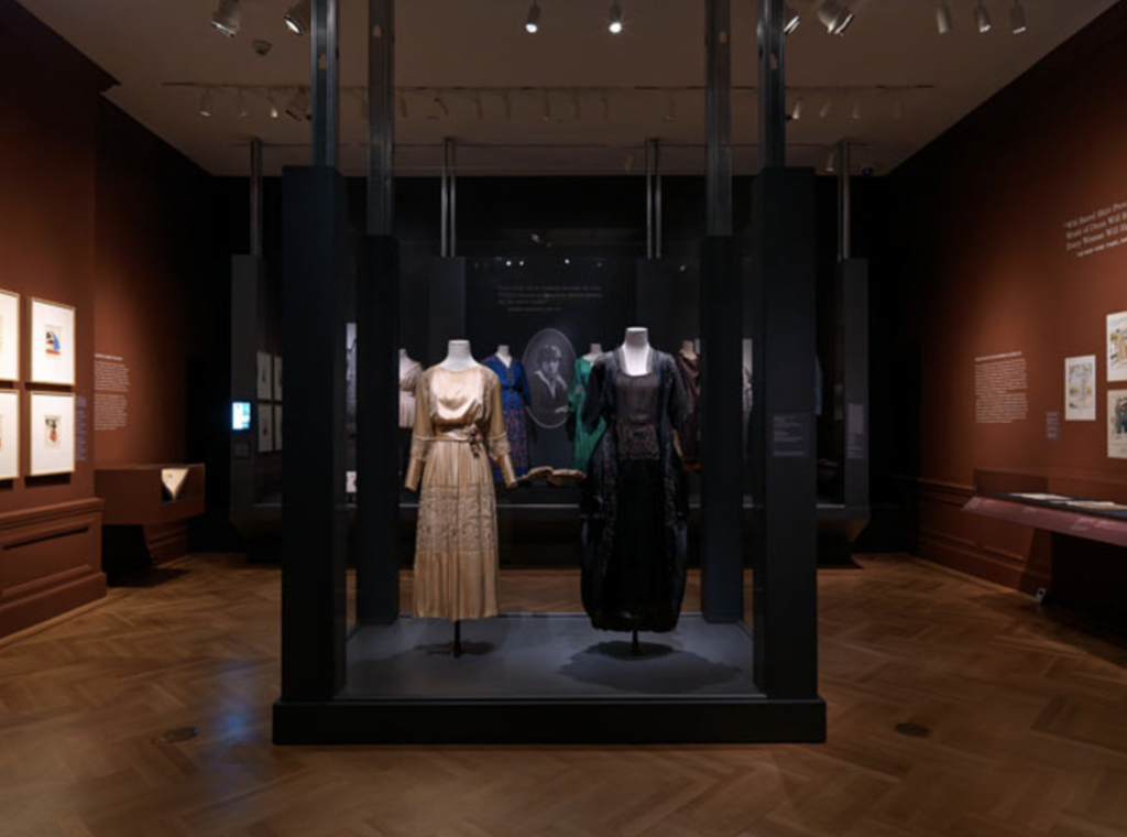 Exhibition with plinth surrounded by glass with two mannequins displaying garments.