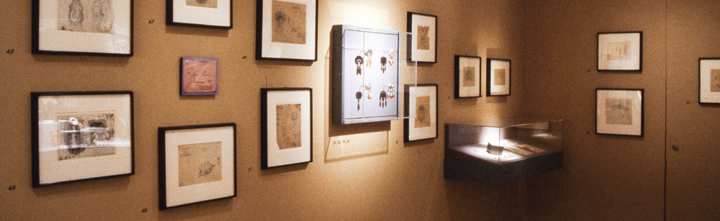 Exhibition with images hanging in frames on the wall, a vertical glass cabinet hanging on the wall displaying jewellery, and a flat cabinet on the wall displaying jewellery.