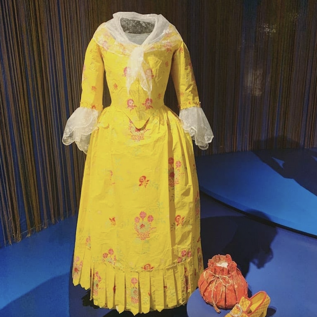 Exhibition with a stripy blue plinth holding a mannequins displaying a yellow dress and smaller plinth holding a red bag and a shoe.