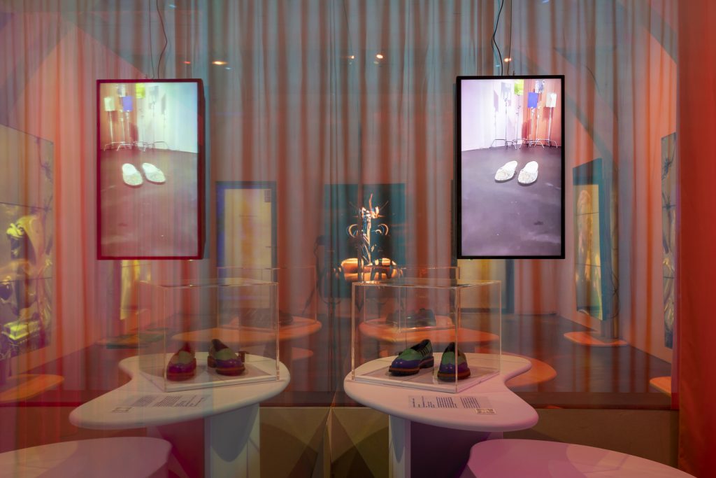 Exhibition display of screens projecting imagery with shoes in the foreground on kidney shaped tables and transparent curtains draped in foreground