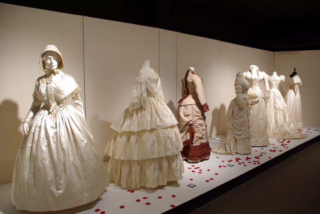 Exhibition with a cream colour wall and a white plinth covered in red rose petal leaves holding mannequins displaying garments.