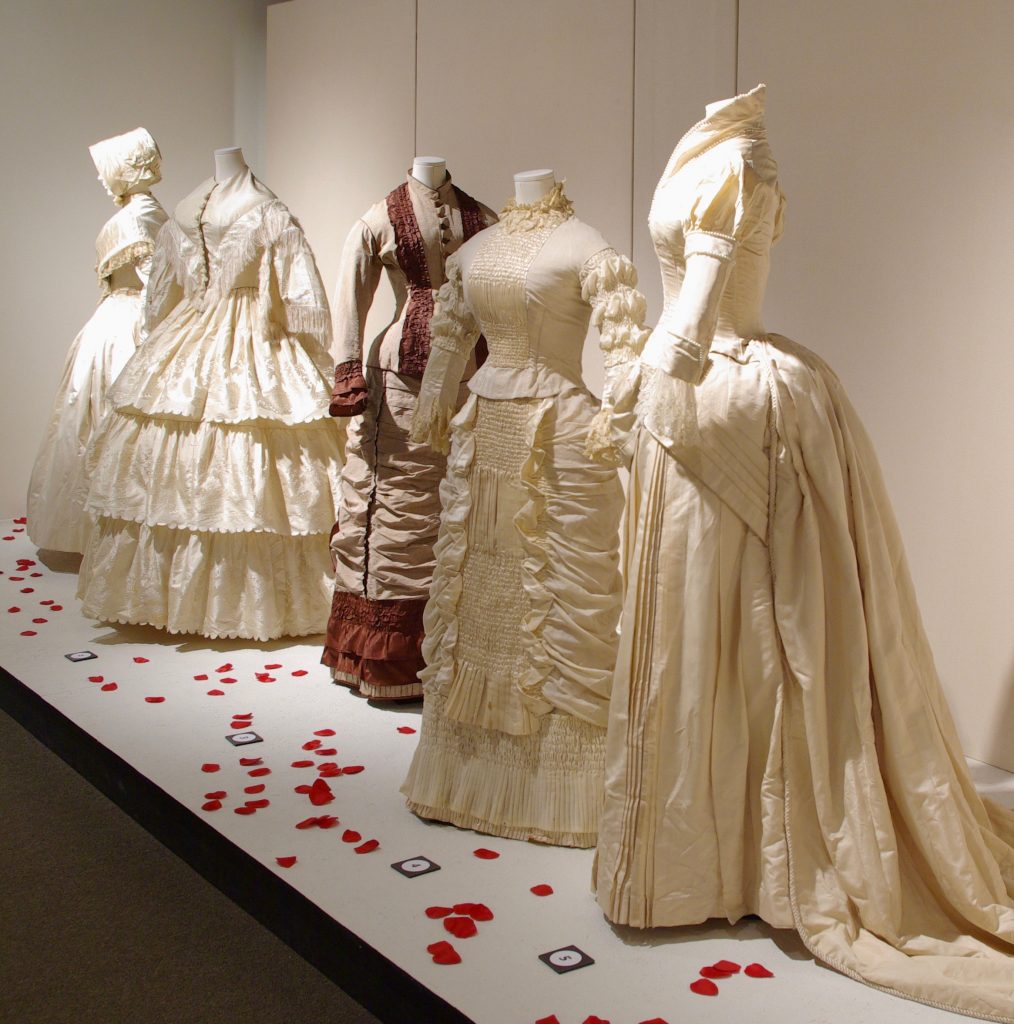 Exhibition with a cream colour wall and a white plinth covered in red rose petal leaves holding mannequins displaying garments.