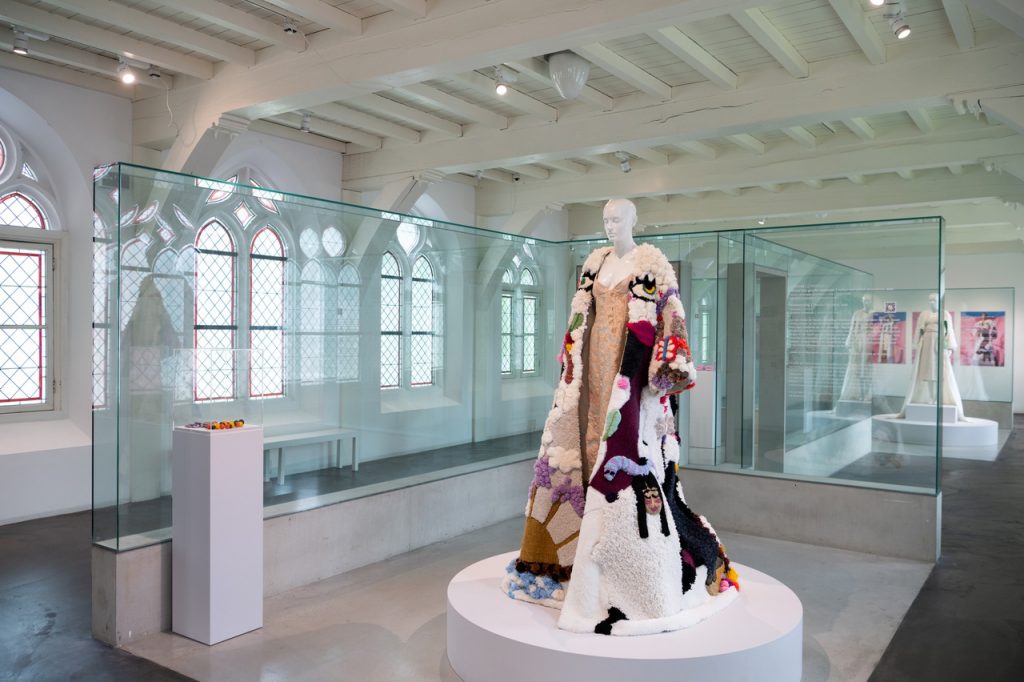 Exhibition installation shot of mannequin with colourful crafted cape in multitude of textures