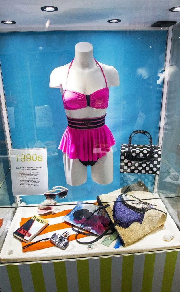 exhibition of mannequin in historic swimwear - pink bikini with a pleated pink skirt, underneath which sit beach accessories of towel, magazine, camera, bags