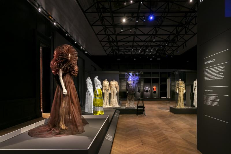 A selection of dresses worn on white mannequins stand on matte black plinths. In the forefront is a brown sheer dress with a layered circular structured design around the shoulders and head.