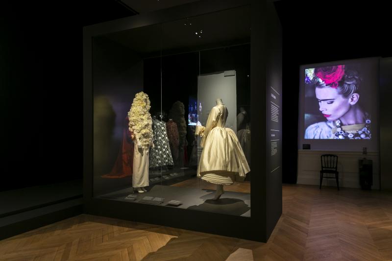 Two cream dresses face each other in a black display case, one with a long blonde wig. In the background is a projection of a model dressed as Frida Kahlo.