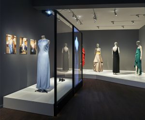 A pale blue sleeveless dress is displayed at the front of the image, displayed right of three photographs showing a model wearing the dress. In the background are four more dresses standing on a wide white plinth.