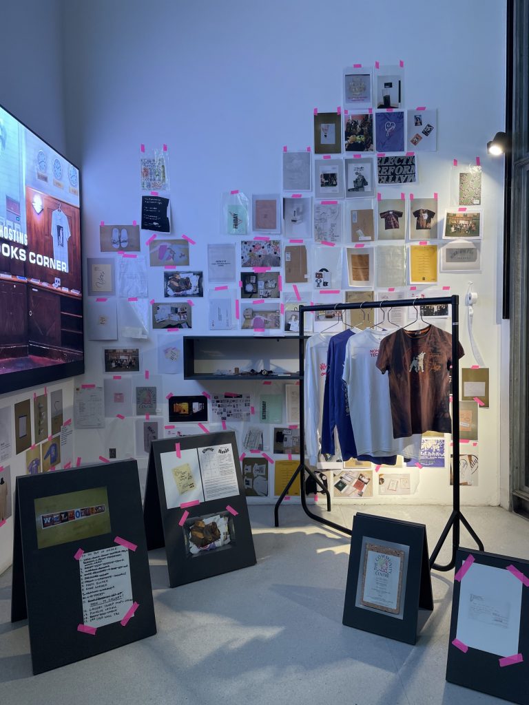 Exhibition display of cards/imagery on wall alongside a flatscreen tv. In the middle sits a clothes rail with garments hanging.