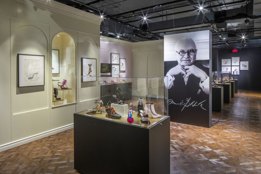 exhibition display of shoes in several glass cases in gallery. To the background is a large black and white photograph of Manolo Blahnik.