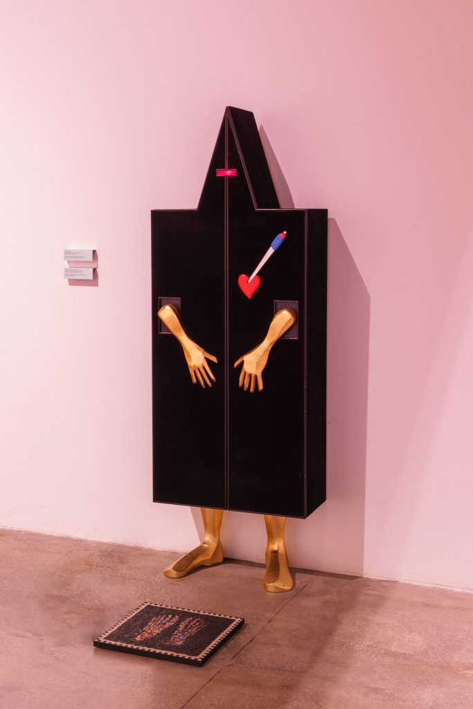 Exhibition display of a black box against a wall housing a mannequin. The box reaches a triangular point at the top and is adorned with gold painted arms and a heart with an arrow through it. mannequin gold ankles, feet visible.