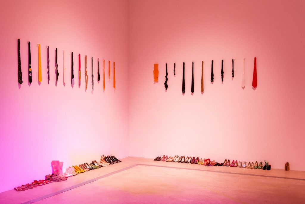 Exhibition installation shot of colourful ties mounted on 2 walls meeting at the corner and colourful pairs of shoes on the floor.
