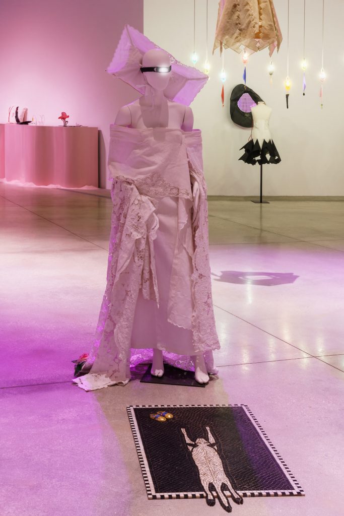 Exhibiiton display of mannequin dressed in white drapes with head dress an head torch. On the floor in front is a doormat depicting a cat stretched out belly up.