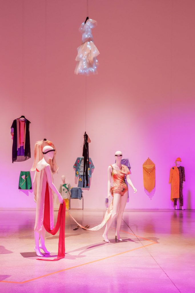 Exhibition installation shot of mannequins with colourful draped fabrics and with colourful garments mounted on wall and from ceiling