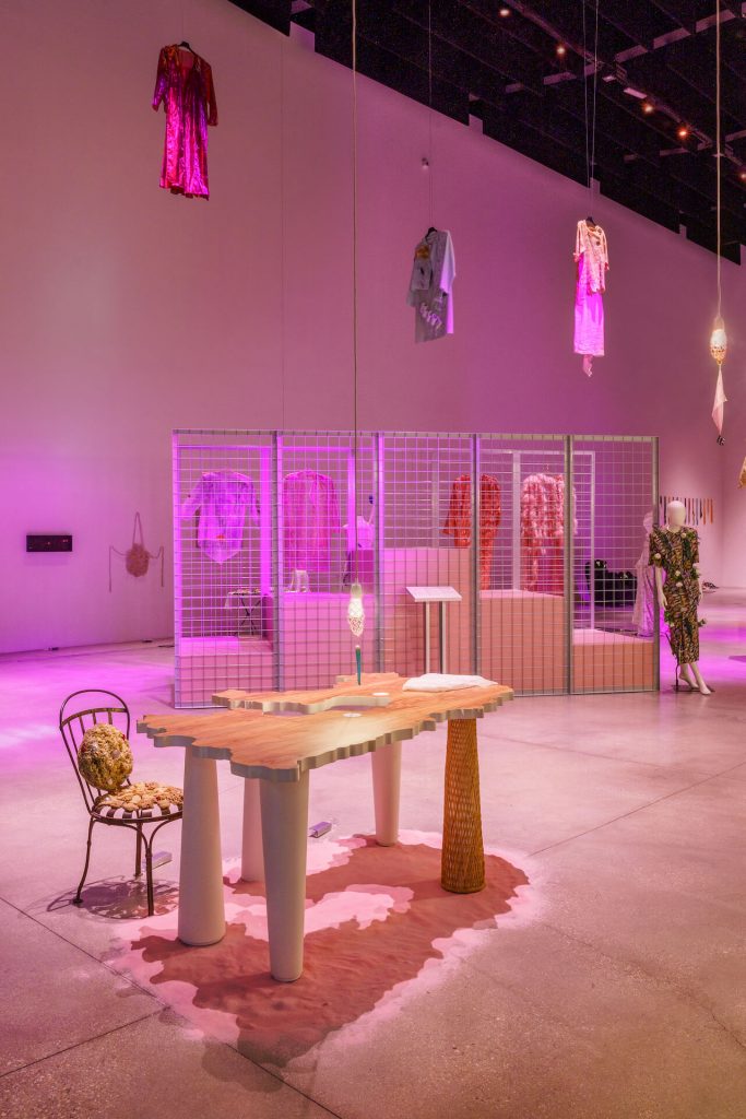 Exhibition display of a carved table to look like a country with thick legs, alternately tapering towards the bottom/top with metal chair and cushions. In the background a metal cage houses hanging garments, and garments are suspended from ceiling. A dressed mannequin is to the right.