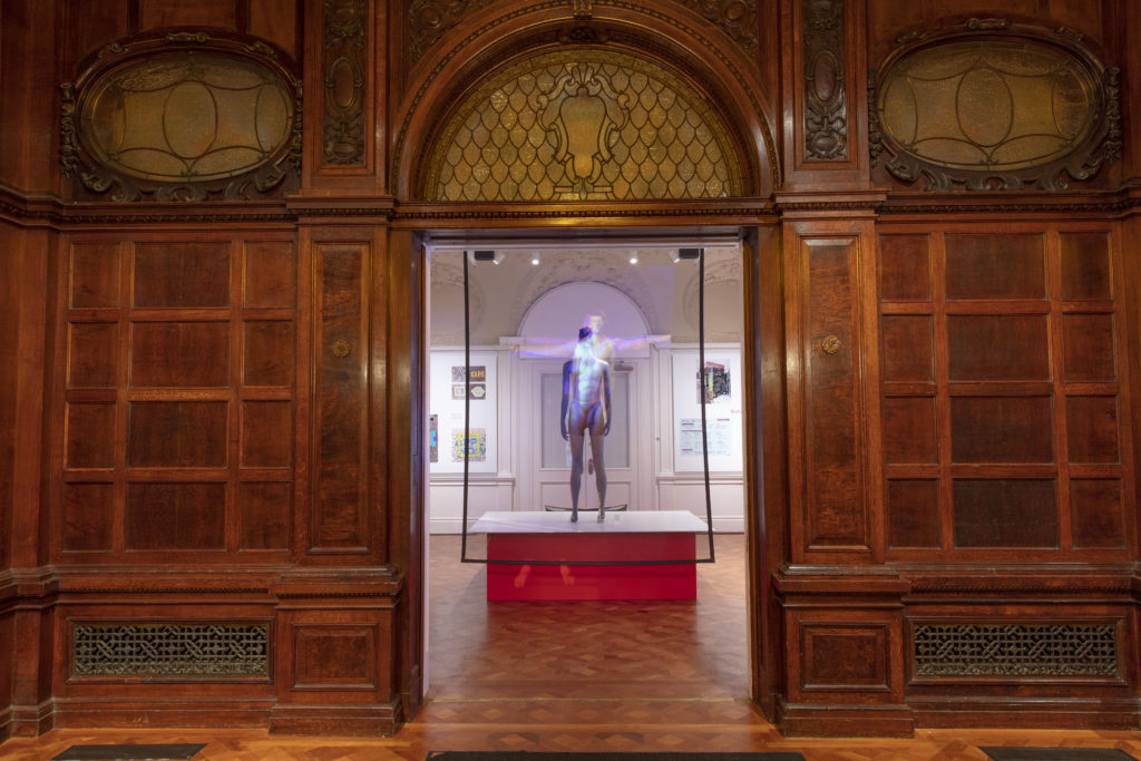 xhibition display of a doorway in wood panelled wall through which can be seen grey male mannequin on plinth in underwear briefs with hologram of outstretched arms.