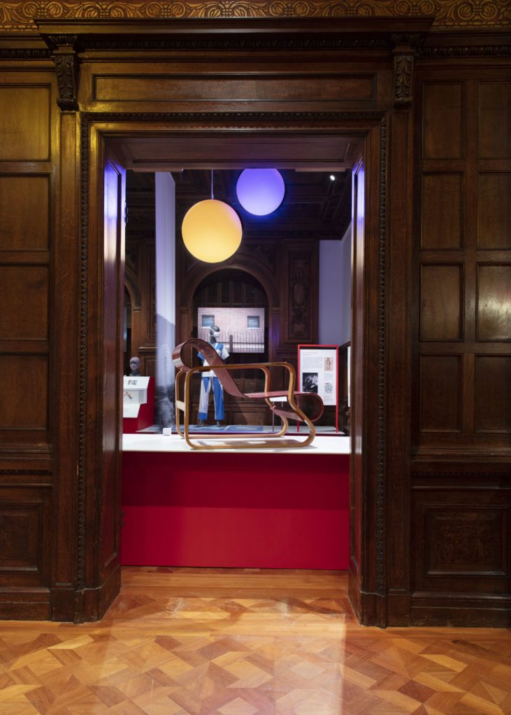 Exhibition display of a doorway in wood panelled wall through which can be seen a wooden curved chair on a plinth.