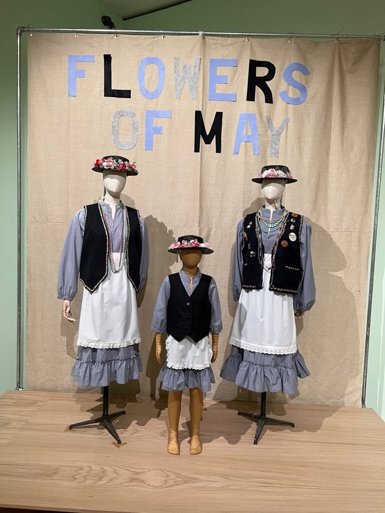 3 Mannequins, 2 adult, 1 child, dressed in May Day outfit of blue dress and hats in front of Flowers of May banner from the Minehead Traditional Sailor's festival