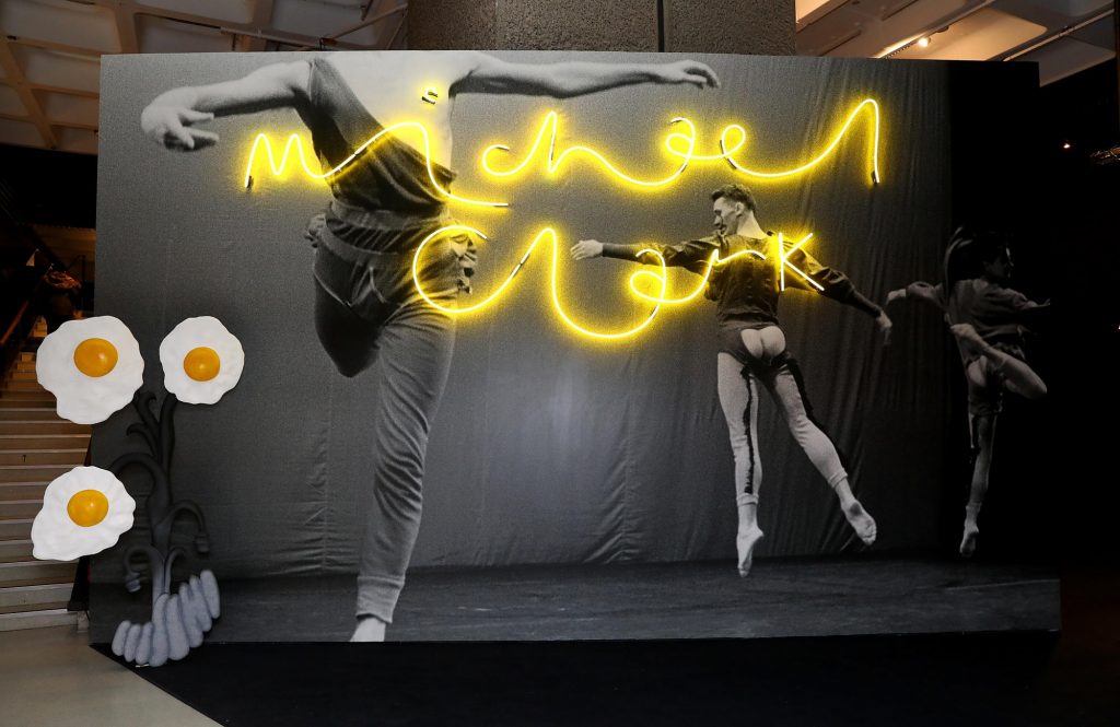 Image of three dancers mid-air and three fried eggs. 'Michael Clark' is written in a yellow neon light.