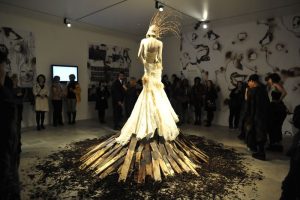 The back of a mannequin wearing an ivory dress and feathered headpiece stands on top of a bonfire in a gallery. The walls of the gallery have scorch marks.