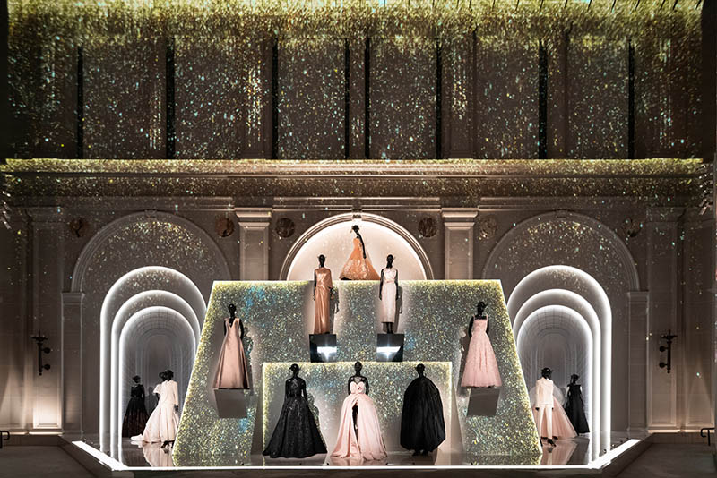 A tiered display of ballgown dresses against a backdrop of projections showing glitter falling