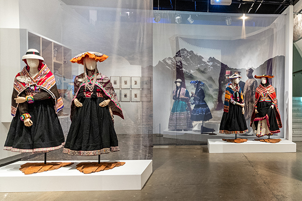 Six Peruvian outfits displayed in pairs on mannequins on white plinths against sheer grey material