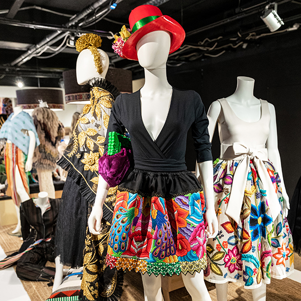 A display of exhibits. In th front is a mannequin wearing a black top with a colourful skirt embroidered flowers and peacock fearthers