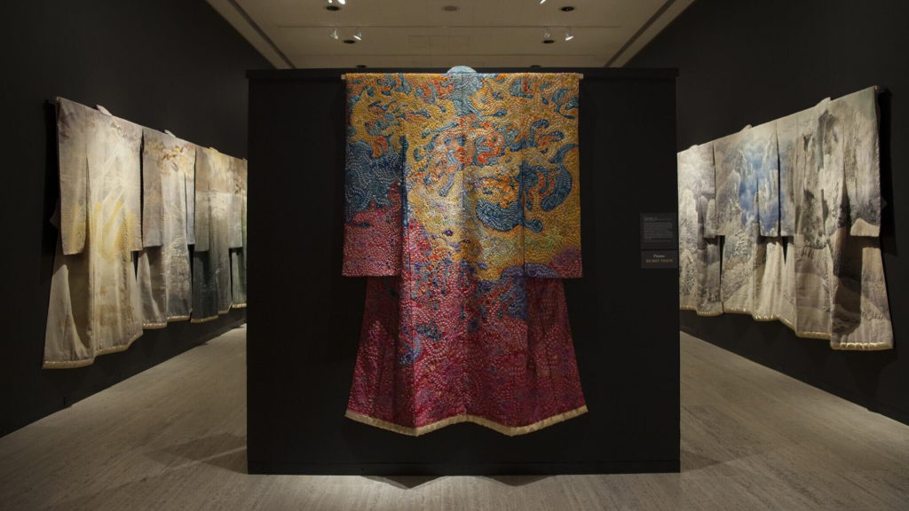 Several pale kimonos hung on two walls. In between the walls is a yellow, blue and red kimono on display.