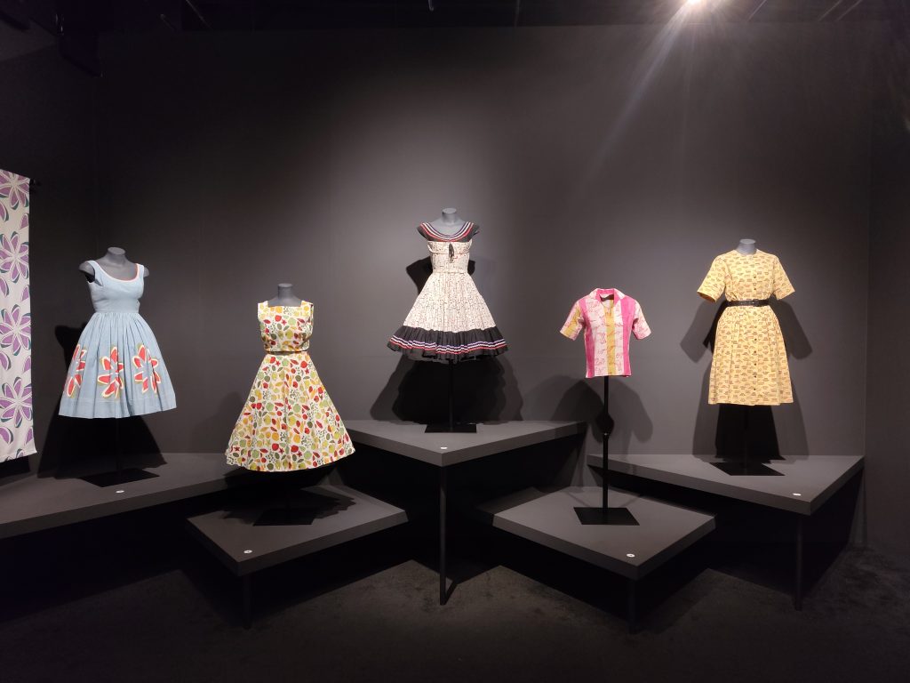 A row of prom-style dresses, short-sleeved shirt and 50s dress on display on tailor's dummies. Each dummy stands on individual square black plinths in front of a black wall.
