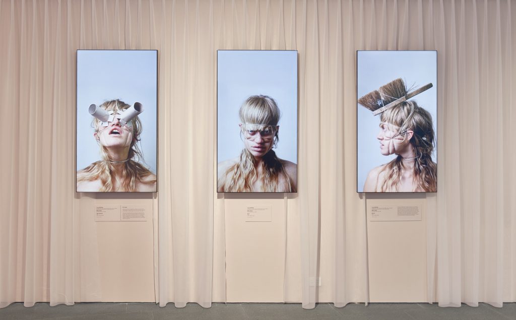 Three exhibition photos of a model wearing every day objects on their face, including toilet rolls and a brush