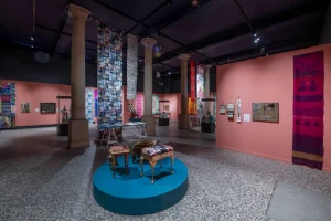 Gallery view of art installations hanging in hues of white and blue. Furniture in hues of brown and black is displayed in the centre.