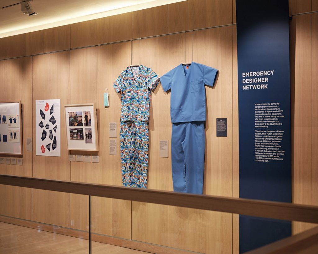 Two sets of hospital scrubs, ine blue and one with a blue, white and yellow pattern, hung alongside a text panel titled 'Emergency Designer Network'