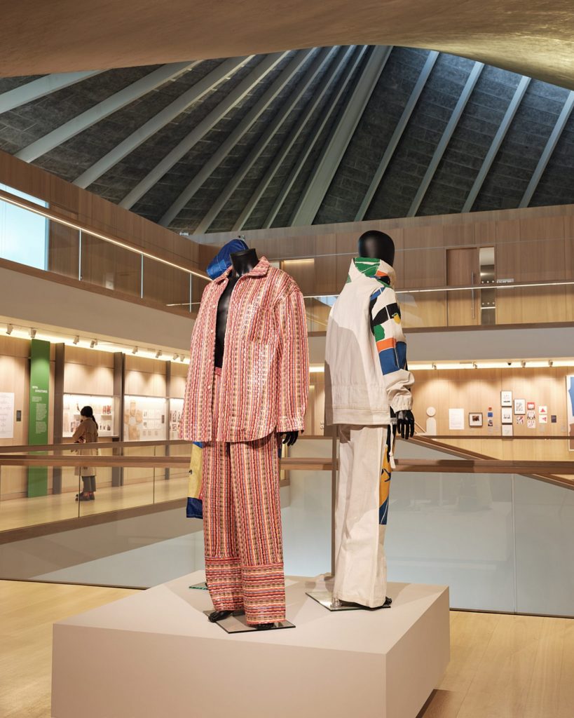 Two outfits displayed on black mannequins. One outfit is a loose open shirt with trousers, both in a striped red pattern. The other outfit, a jacket and trousers, has a cream back with blocks of orange, green, blue and back blocks on the sleeves.