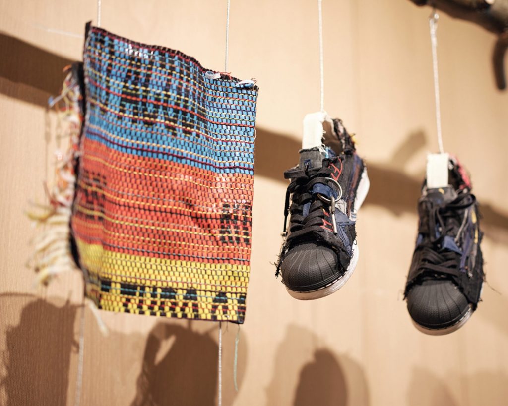A pair of black shell-toe trainers are hung by white rope alongside a woven blue, orange and yellow piece of fabric with some text printed on it. The fabric of the trainers are ditsressed; one is laced and the other is not
