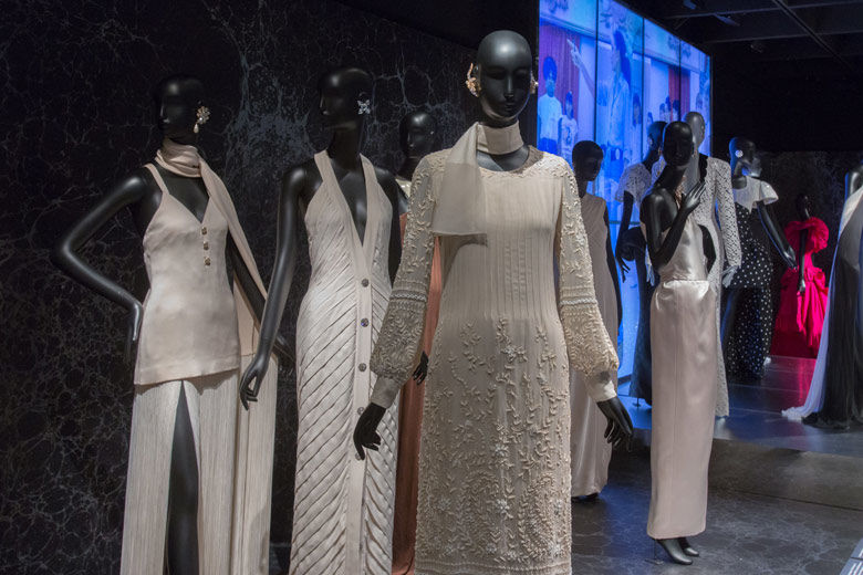 Three silvery-grey outfits are worn on mannequins on the foreground. More outfits are shown in the background, in front of a large screen showing a runway show.