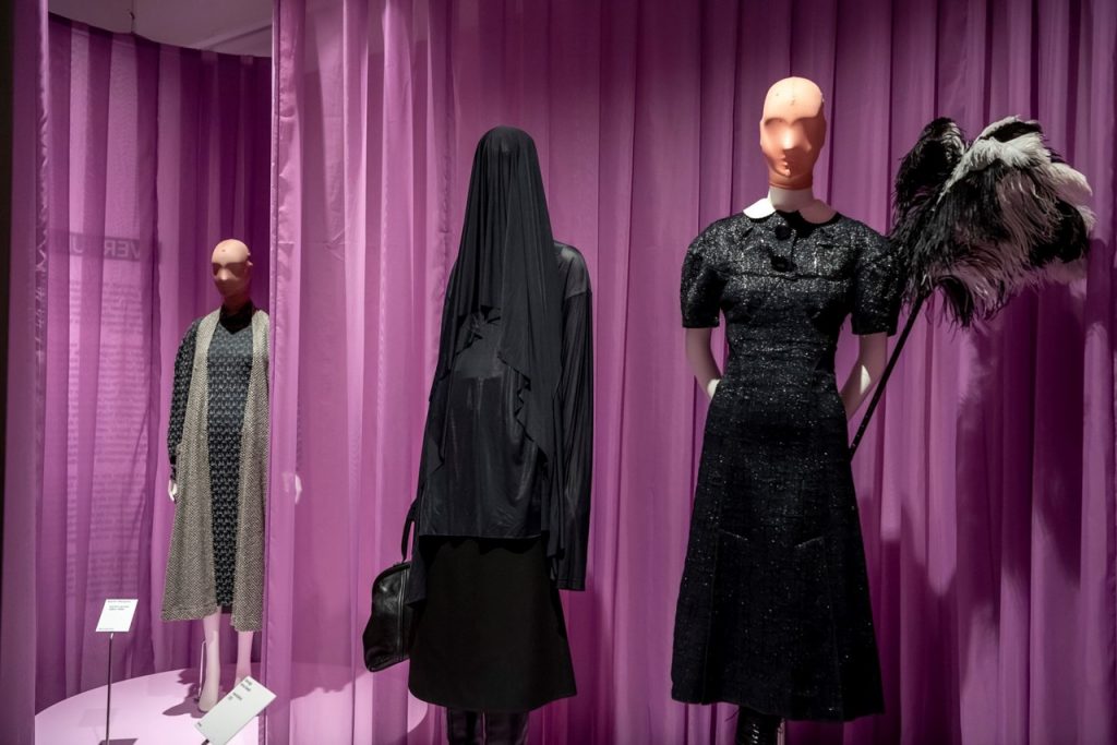 Three black outfits on mannequins are set against purple material dividing the space. Behind the mannequin on the right is a black and white duster with plumes of feathers.