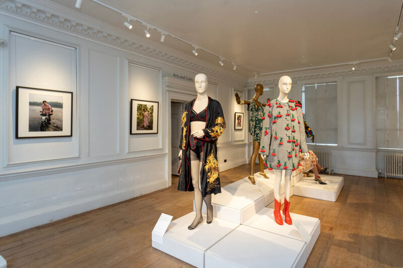 Four mannequins, including a dress with appliquéed cherries and black underwear, stand in a central plinth, surrounded by photographs